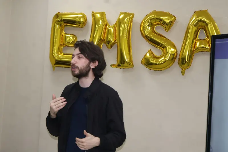 A speaker addresses the room in front of the gold EMSA balloons